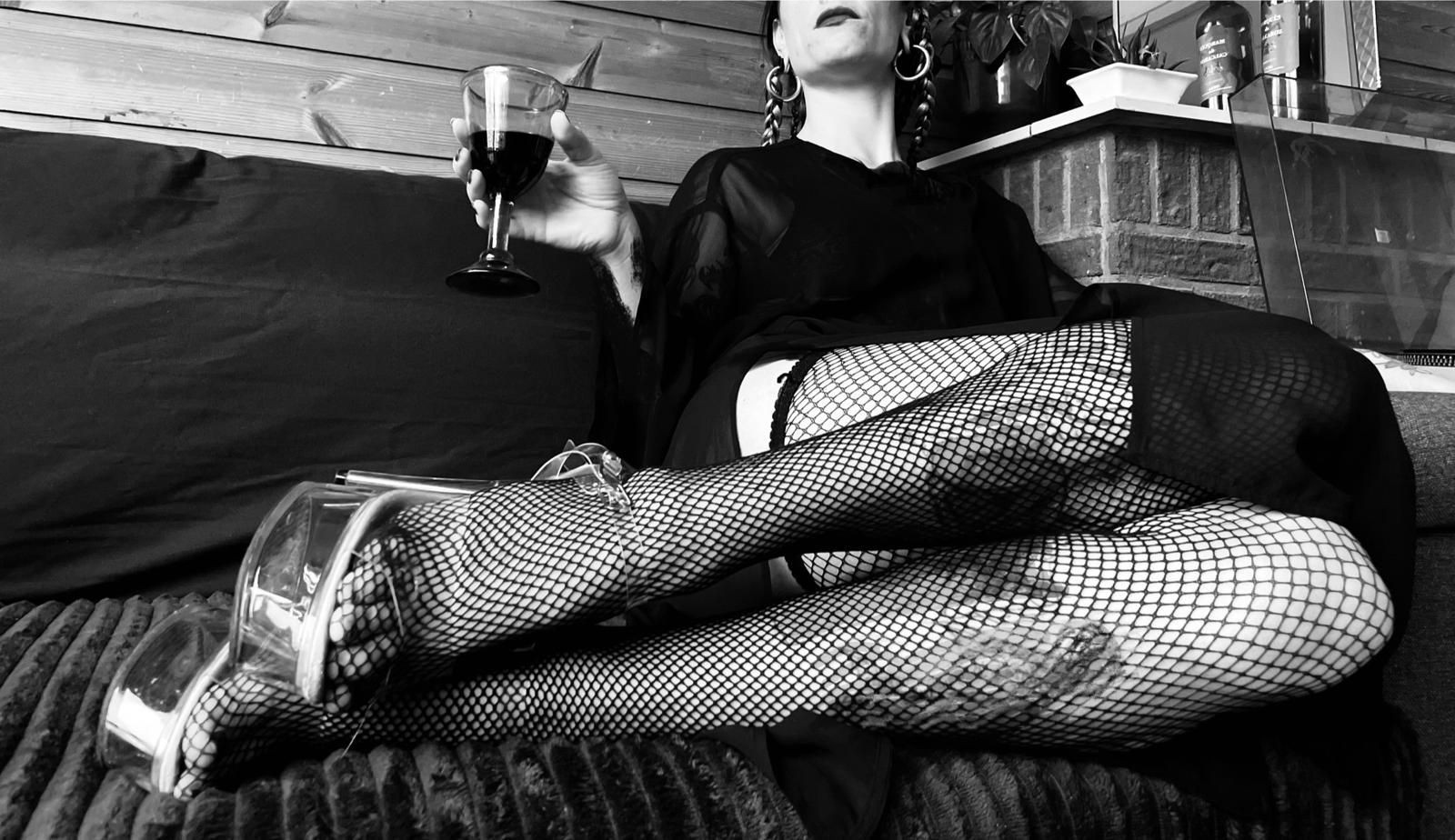 Woman in fishnet stockings and high heels holding a glass of wine on a sofa.